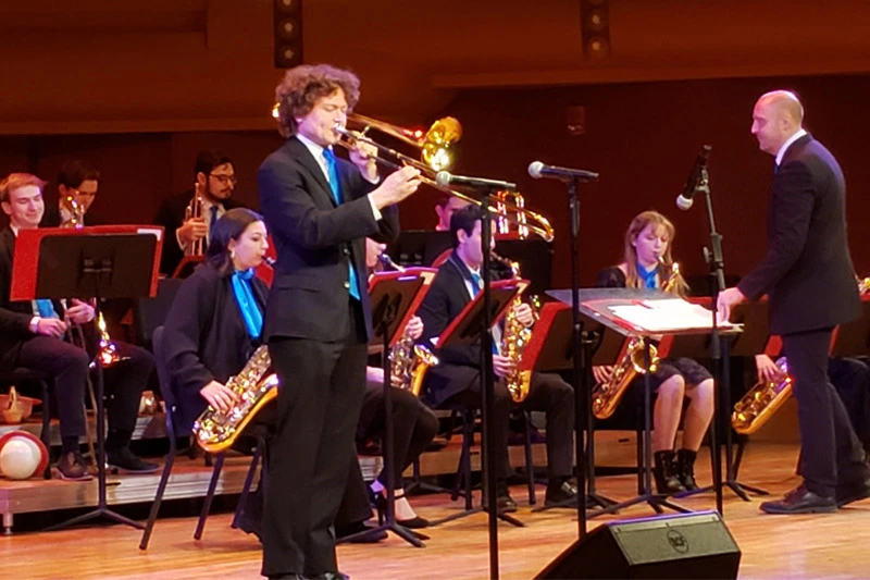 A male trombonist plays onstage with a jazz band, wearing a dark suit with a bright blue tie. Other musicians sit behind red music stands, playing saxophones and other brass instruments. A conductor in a dark suit stands to the right, following the score.