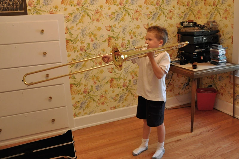 Young boy in a white shirt and dark shorts plays a gold trombone in a room with floral wallpaper.