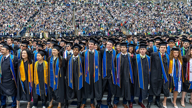 Graduates in black gowns and blue and gold stoles stand together in Notre Dame Stadium during Commencement.