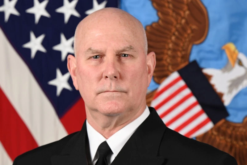 A bald man in a dark suit and white collared shirt and tie faces forward. He has a serious expression. The U.S. flag and the Joint Chiefs of Staff flag are visible in the background.