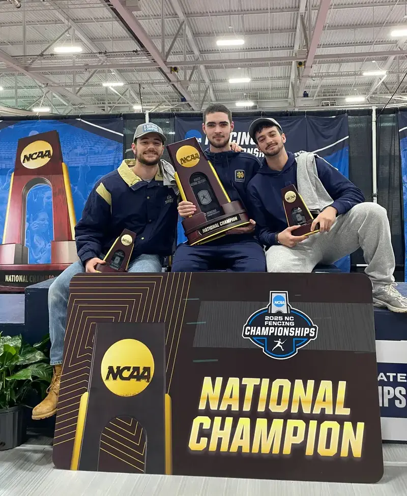 Three Notre Dame fencers pose with their NCAA National Championship trophies. They sit on a platform in front of signage for the 2025 NCAA Fencing Championships.