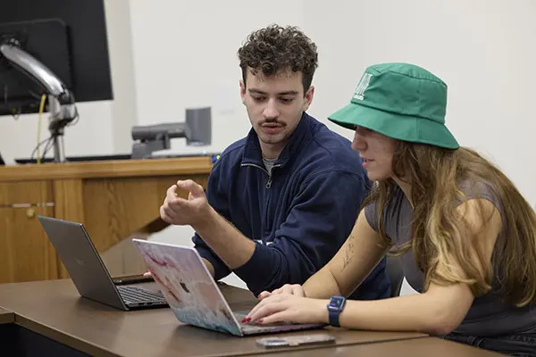 Two students collaborate on a laptop in a classroom. One student, wearing a navy blue jacket, gestures while speaking. The other student, wearing a green bucket hat, types on a decorated laptop.