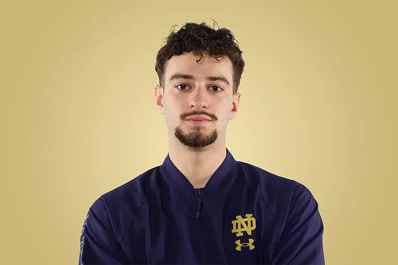 Headshot of a male with short curly hair and a goatee, wearing a navy blue jacket with a gold Notre Dame monogram and Under Armour logo against a gold background.