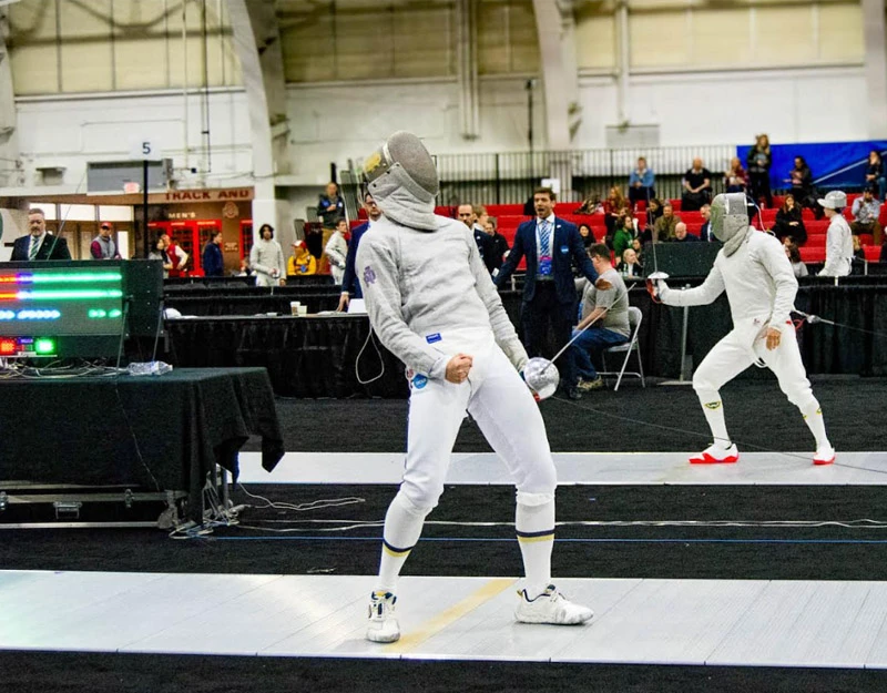A fencer in white and gold uniform celebrates a point, fist clenched, during a match. 