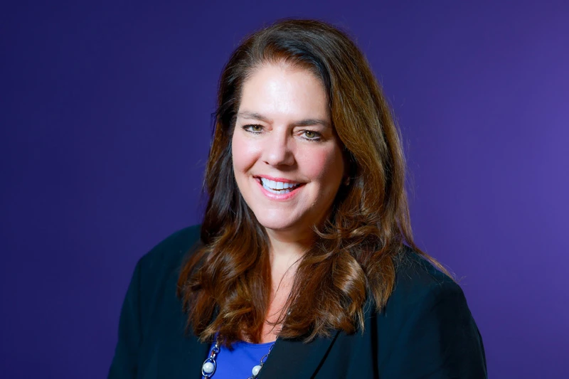 A white woman with shoulder-length brown hair smiles against a purple backdrop. She wears a black blazer over a royal blue top and a silver necklace.