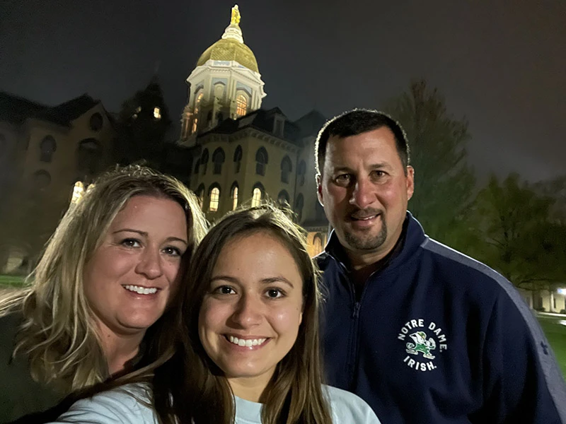 At night, a family of three smiles for a selfie in front of the illuminated Main Building at the University of Notre Dame. The father wears a Notre Dame Irish jacket.