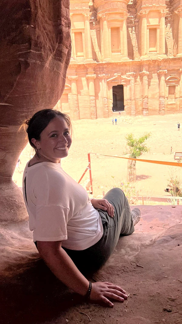 Smiling female in a white shirt and greenish-brown pants sits inside a rock alcove, looking back over their shoulder at Ad Deir (The Monastery) at Petra, Jordan.