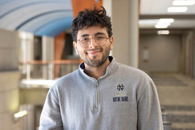A male student wearing a gray Notre Dame quarter-zip smiles in a campus building hallway.