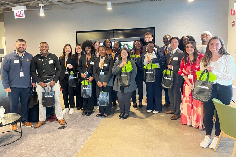 A group of students and professionals pose for a photo holding Deloitte tote bags.