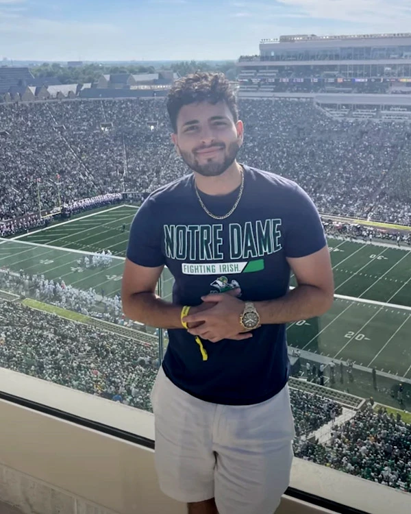A person wearing a navy blue 'Notre Dame Fighting Irish' t-shirt and white shorts stands with hands clasped in front of them, overlooking Notre Dame Stadium filled with a crowd on a sunny day.