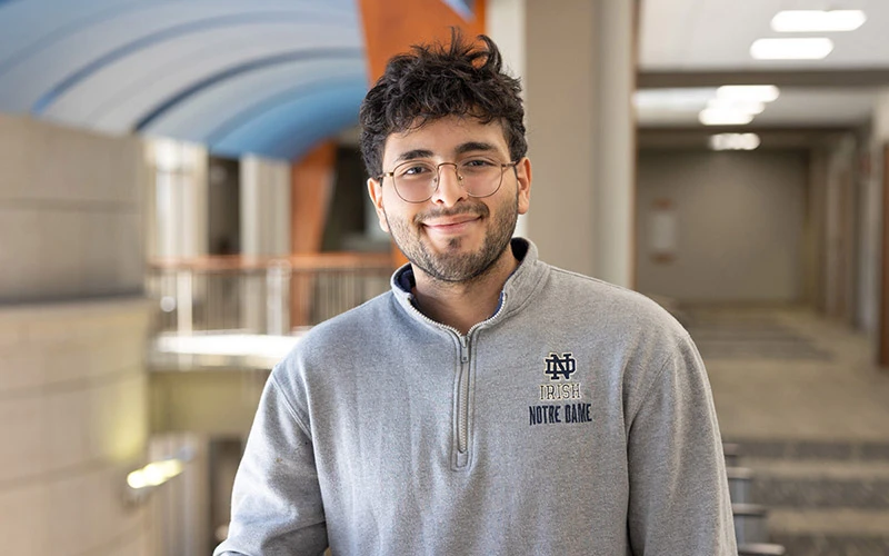 A male student wearing a gray Notre Dame quarter-zip smiles in a campus hallway.