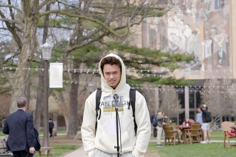 A student wearing a white hooded sweatshirt stands at a microphone on campus with the Hesburgh Library mural in the background.