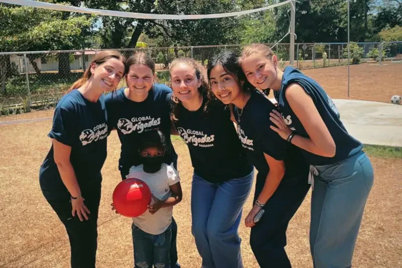 Six Notre Dame students in 'Global Brigades Panama' t-shirts pose with a young child holding a red ball on a dirt court with a volleyball net.