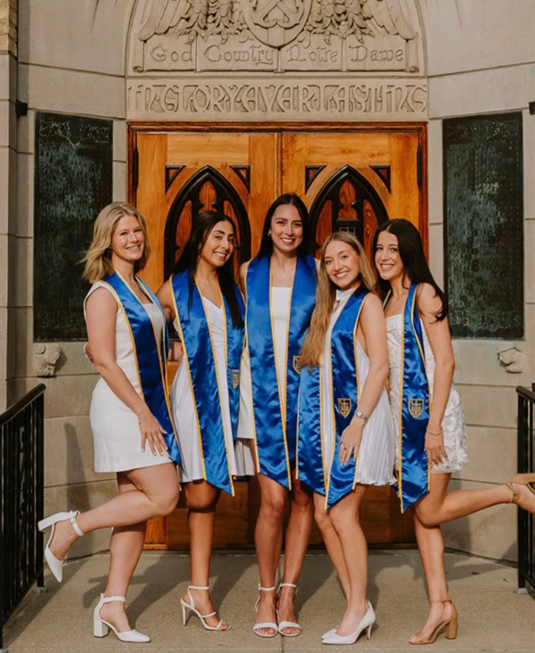 Five women in white dresses and blue and gold stoles pose in front of a doorway at the University of Notre Dame.