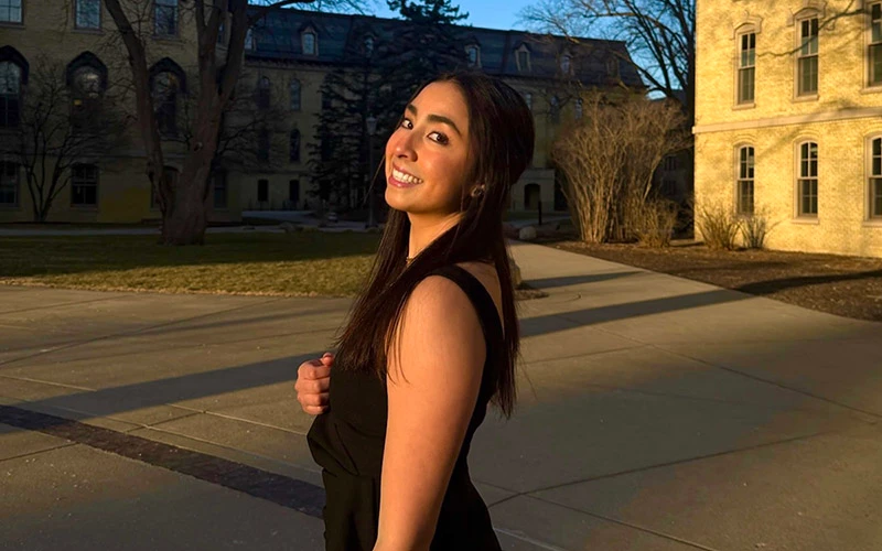 A female student with long brown hair, wearing a black sleeveless dress, smiles over her shoulder on a sidewalk in front of a Notre Dame residence hall bathed in the golden light of late afternoon.