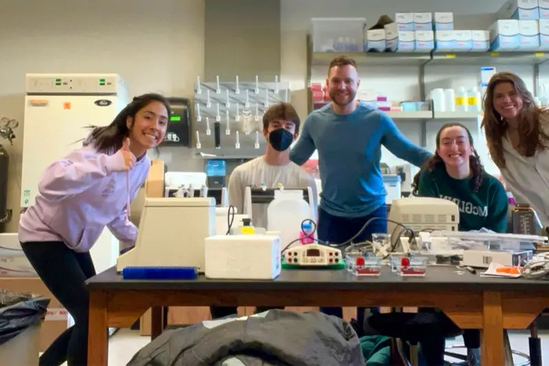 Five students and a professor gather around a lab table with equipment in a science lab.
