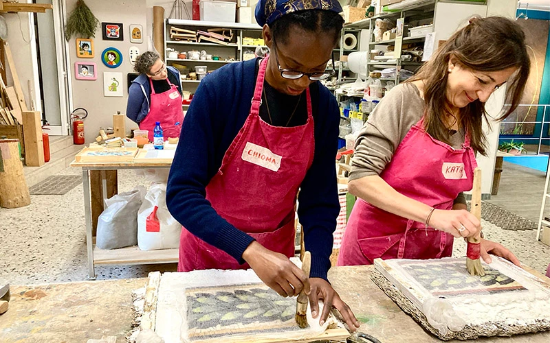 Two people wearing pink aprons use small brushes to apply adhesive to mosaic artwork in an art studio. A third person in a pink apron stands at a workbench in the background.