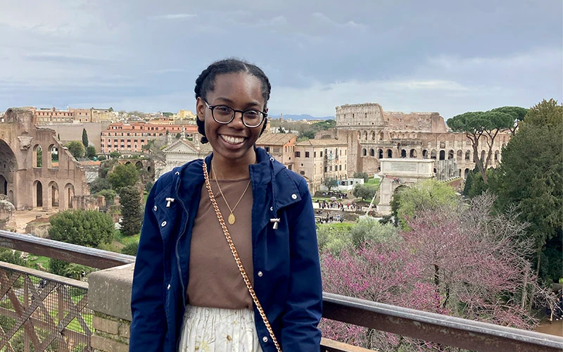 A black female student wearing a blue jacket and glasses stands before a scenic overlook of the Roman Forum and the Colosseum.