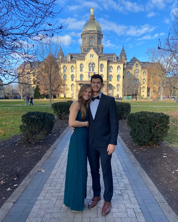 A couple dressed for a formal event poses on a brick walkway in front of the Main Building at the University of Notre Dame. The woman wears a long, teal gown, and the man wears a dark suit with a bow tie. The Golden Dome is visible in the background under a blue sky.