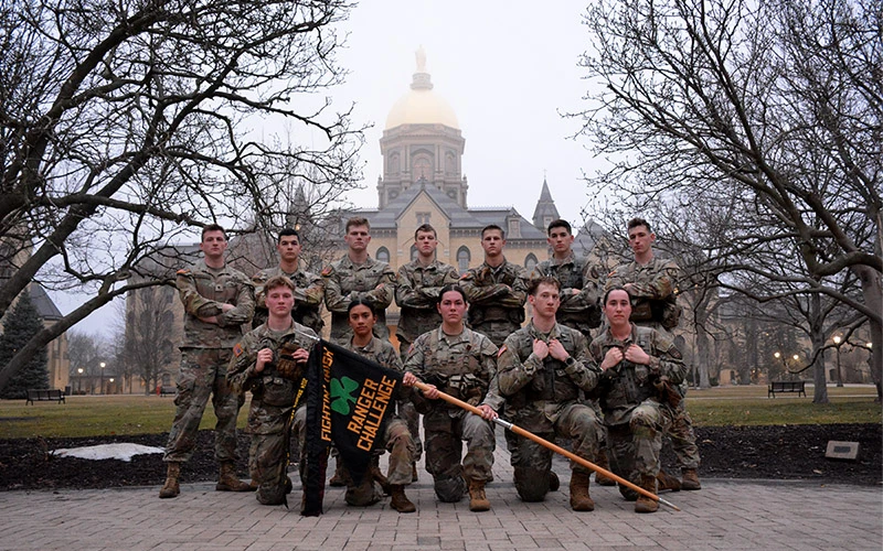 A smiling person in a U.S. Army dress uniform stands in front of an ornate wooden door.