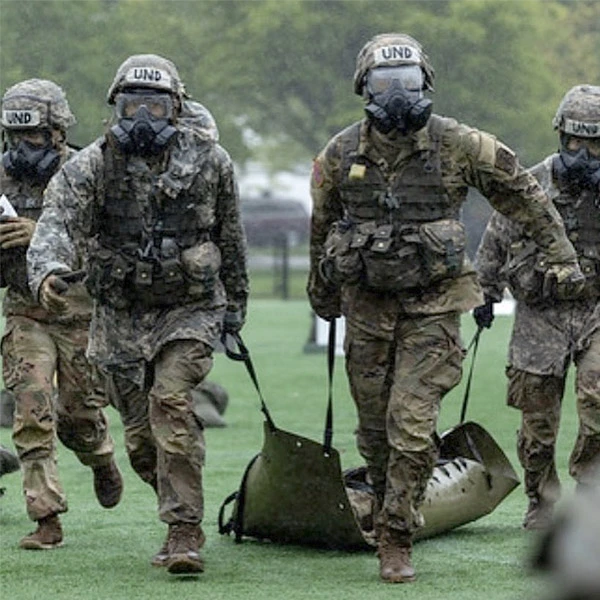 ROTC cadets in camouflage uniforms and gas masks run across a field carrying a stretcher.