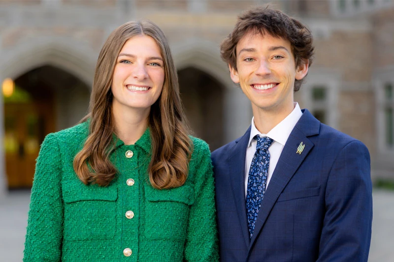 Two smiling students stand in front of an arched entryway of a Notre Dame building. The woman on the left wears a green jacket, and the man on the right wears a navy suit with a floral tie.
