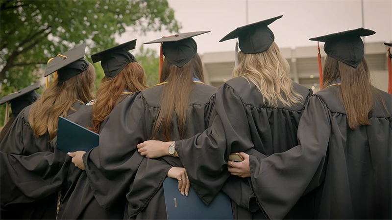 Graduates in black gowns and mortarboards with gold and red tassels stand with arms around each other, some holding diplomas.