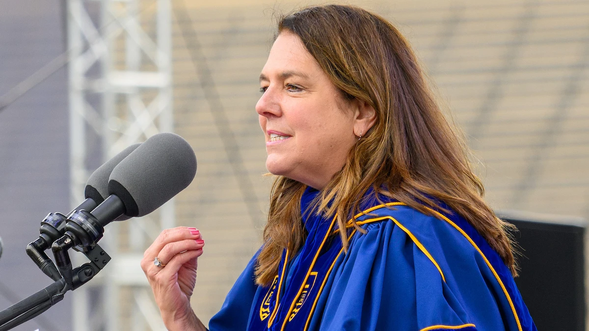 A woman with shoulder-length brown hair, wearing a blue and gold academic robe, speaks at a microphone.