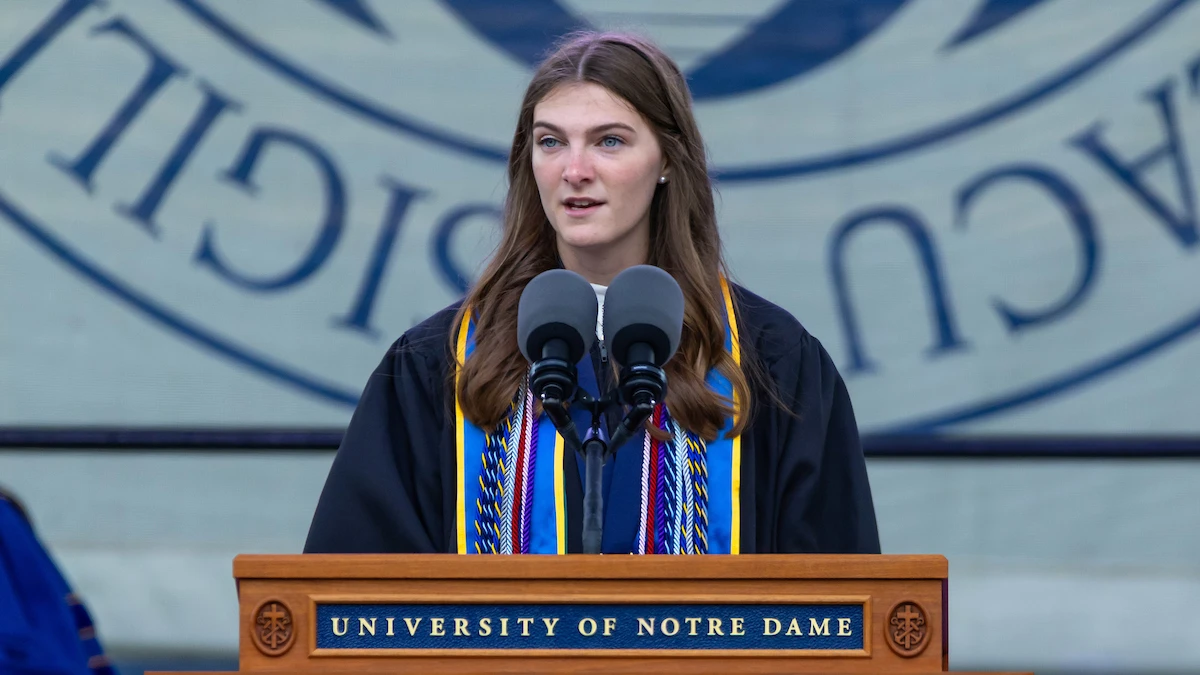 A graduating student with long brown hair, wearing a black graduation robe and colorful cords, speaks at a podium bearing the University of Notre Dame logo.