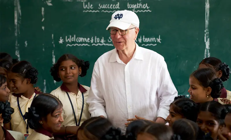 A white man in a white Notre Dame baseball cap and shirt smiles at a classroom of young Indian girls in uniform in front of a chalkboard that reads We succeed together and Welcome to [Symbol] team.