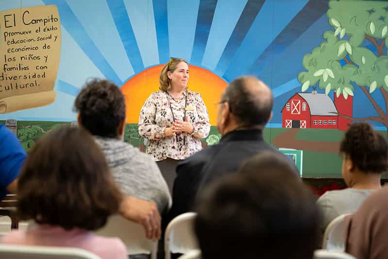 A woman stands in front of a group of people and welcomes them. There's a colorful painting of a farm behind her on the wall.
