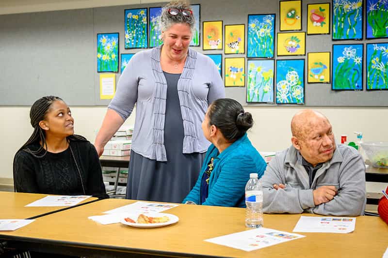 A woman standing talks to two people sitting at a table.