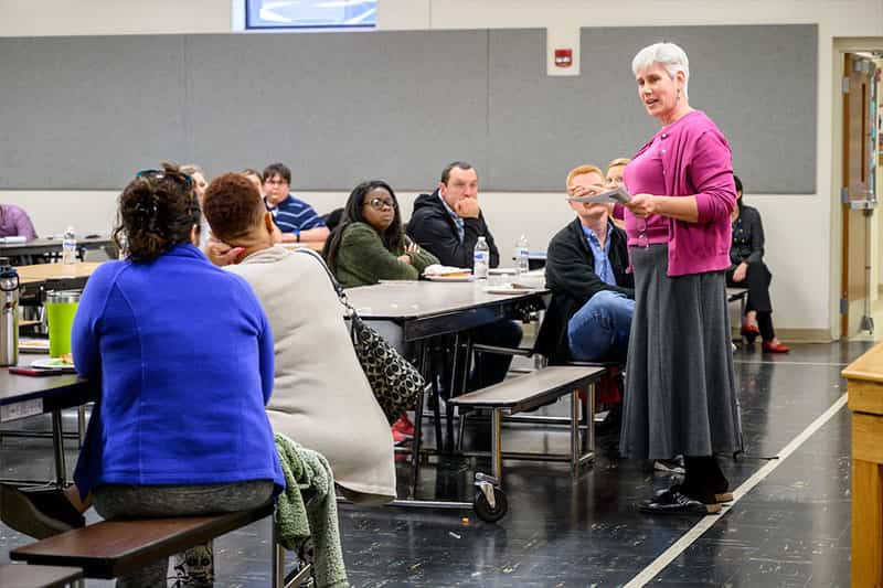 A woman stands in front of a class speaking.