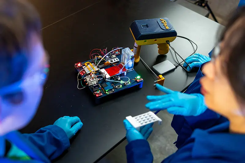 Grad students stand in the blurred foreground with the 'sniffer' device sits on a table in focus.
