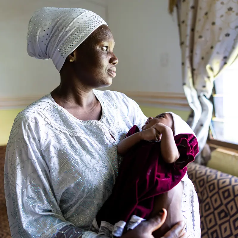 A new mother in a white headscarf and ornate dress holds a baby wrapped in a red and white blanket.