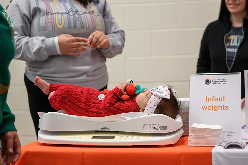A baby in a red knitted outfit and polka-dot headband is weighed on a scale at an information table.