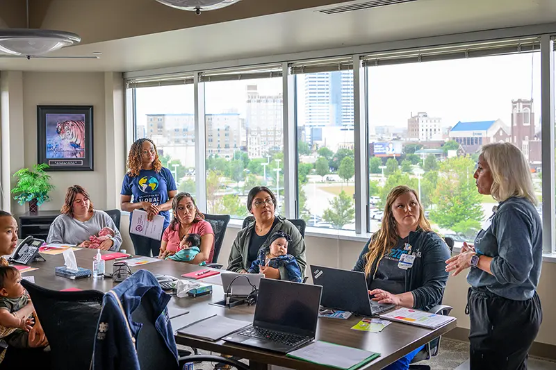 A group of mothers holding babies listens to a speaker in a Beacon Health conference room with a large window overlooking the city of South Bend, Indiana.