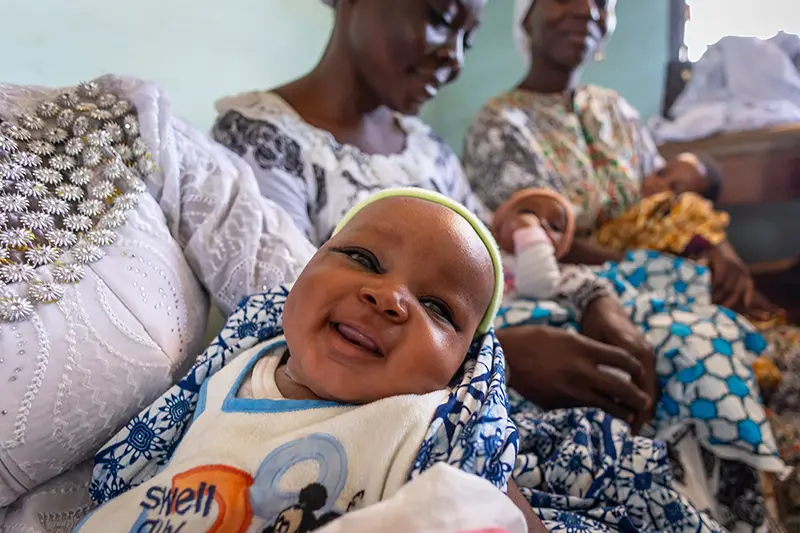 A smiling baby in a bib and patterned blanket is held by a mother seated with other women.
