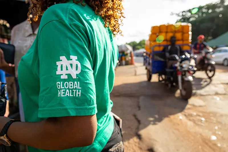 Joyce Adams in a Notre Dame Global Health green shirt stands on a busy market street in Ghana with a motorcycle carrying stacked goods.