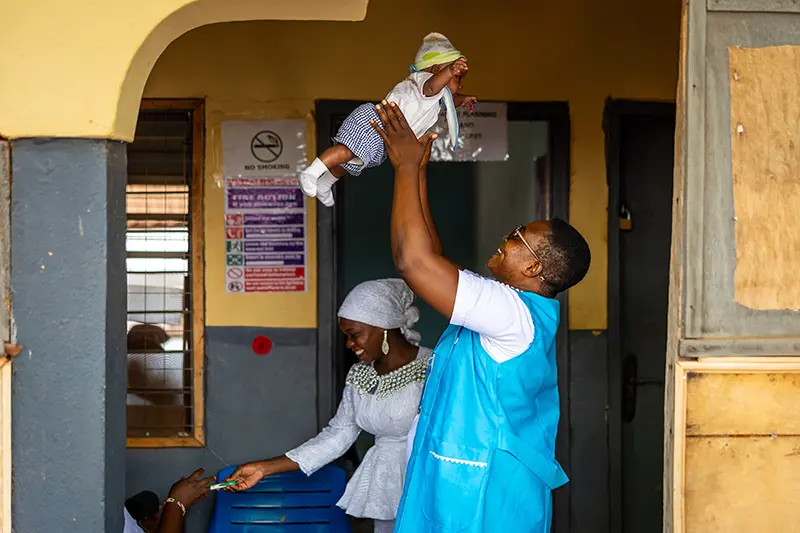 A smiling midwife in a blue vest holds a baby high above their head outside a clinic.