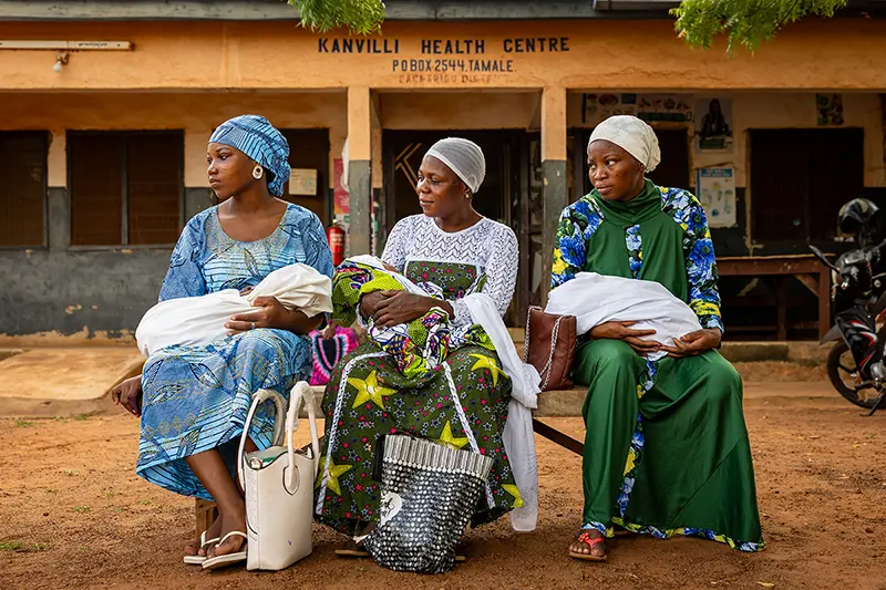 Three mothers wearing traditional clothes and headscarves sit on a bench outside the Kanvilli Health Centre holding babies.