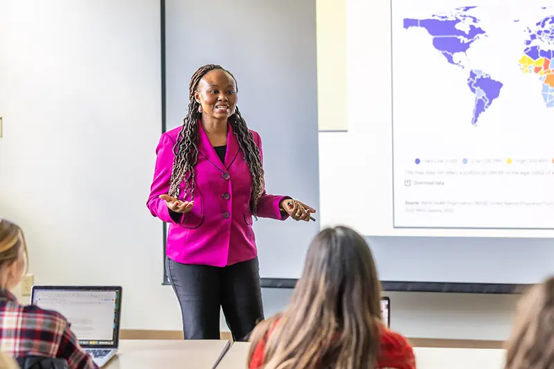 A woman in a vibrant pink blazer lectures using a world map projection to a class of students in O'Shaughnessy Hall.