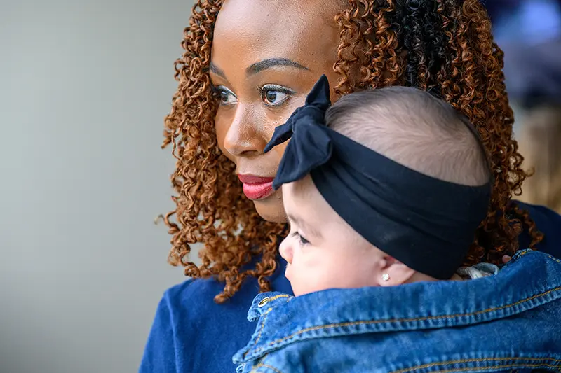 A woman with brown curly hair and red lipstick holds a baby wearing a denim jacket and black bow headband.