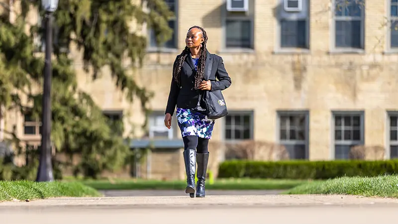 A woman in a black blazer, floral dress, and tall black boots walks on a path in front of a tan brick campus building.