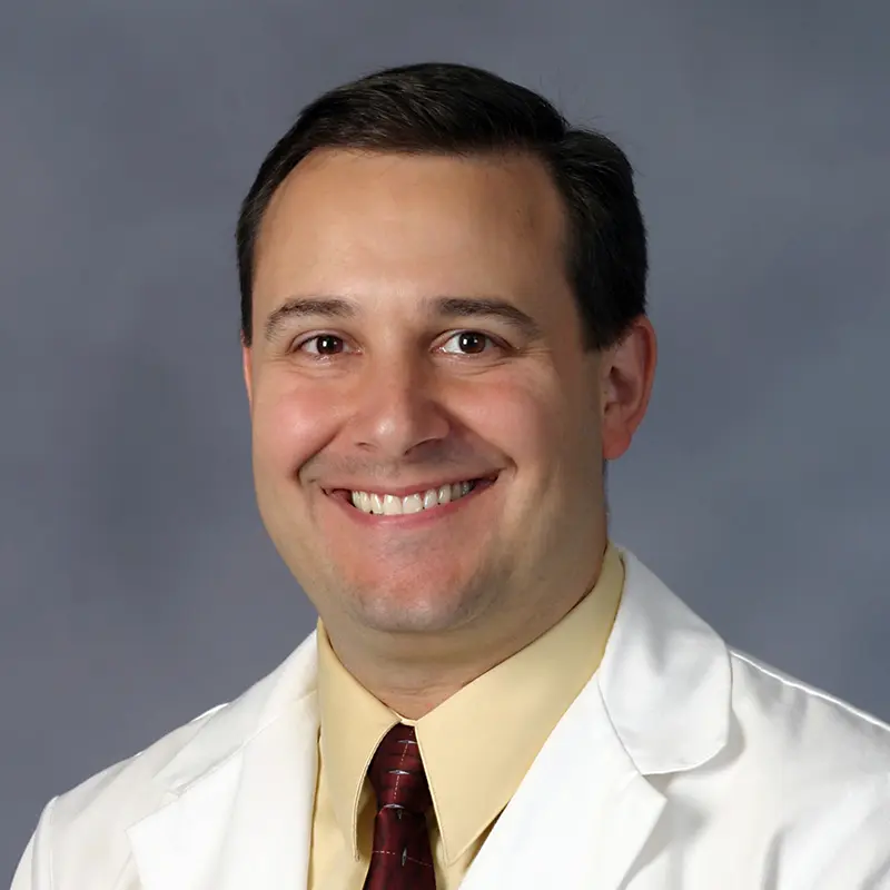 A man in a white lab coat, yellow shirt, and burgundy tie smiles in a professional headshot against a gray background.
