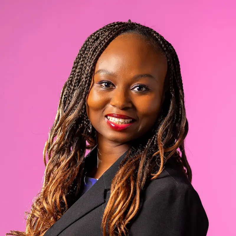 A professional headshot of a smiling woman with long brown and blonde braids in front of a pink background.
