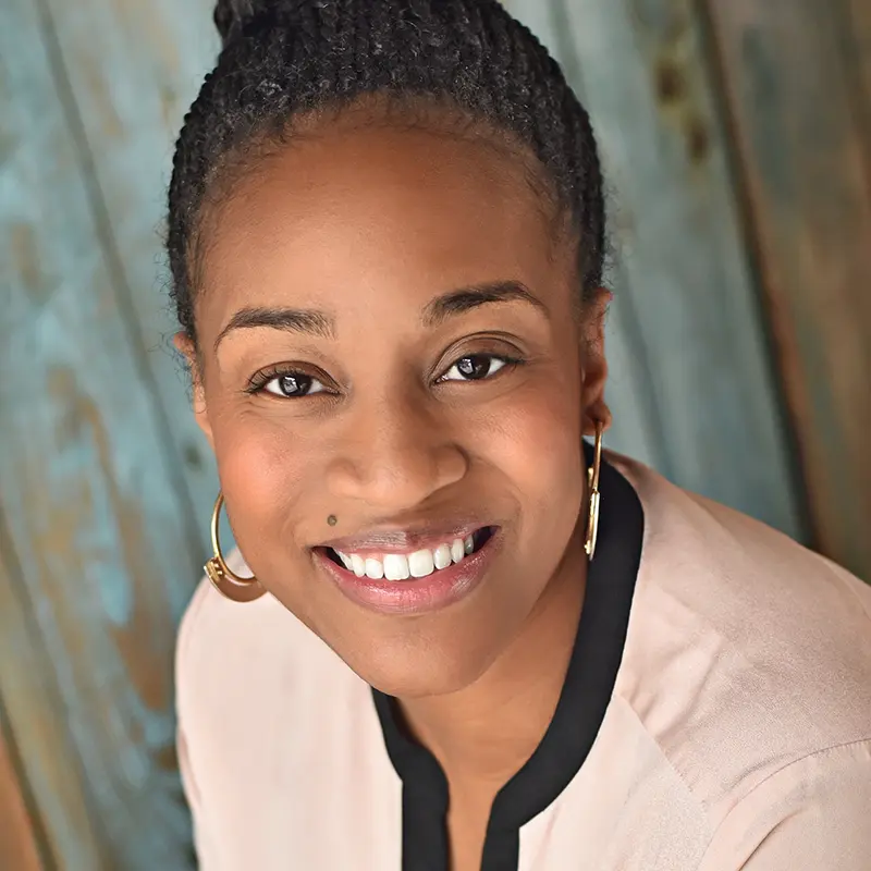 A professional headshot of a smiling woman with her hair in a bun, wearing a light-colored blouse and gold hoop earrings.