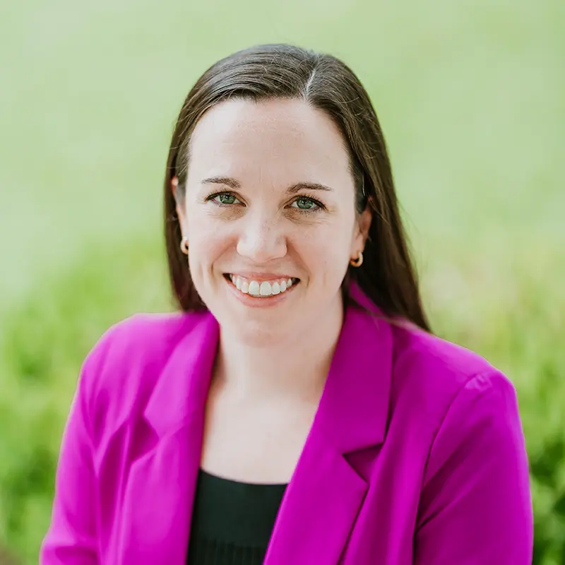 A professional headshot of a smiling woman with long brown hair wearing a bright magenta blazer.