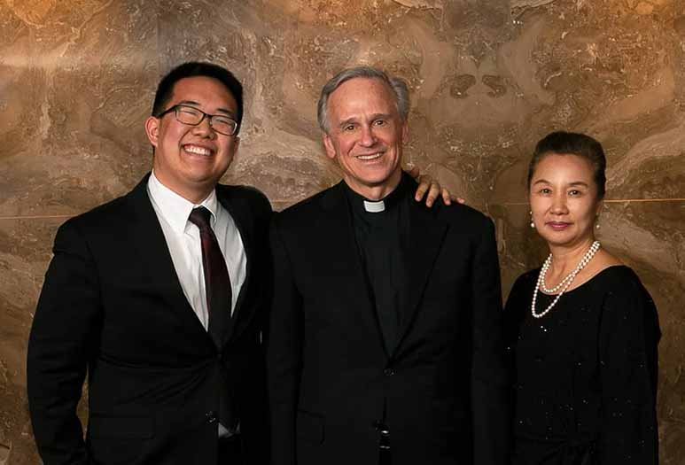 Eric Kim with his mother and Rev. John I. Jenkins, C.S.C. stand in front of a backdrop in a studio.