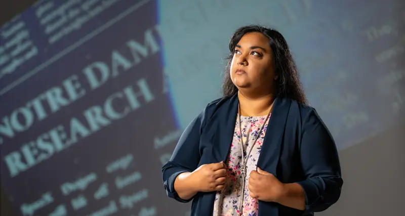 A dark-skinned woman in a blazer and floral top looks upward with 'Notre Dame Research' projected behind her.
